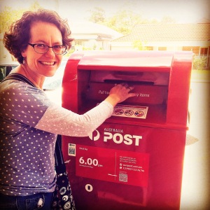 Photo of Rebecca Newman in a spotted shirt posting a letter into a red post box. 